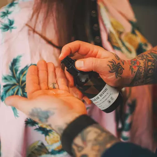 Manos con tatuajes aplicando SHEAR REVIVAL Progress aceite multiusos